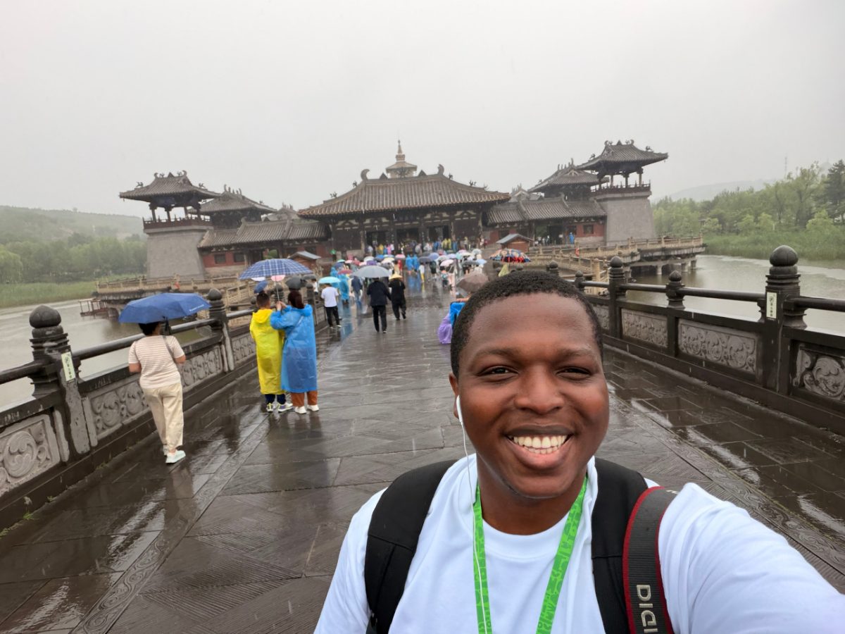 Pamimo smiling in the rain on a stone bridge leading to a temple, with visitors carrying umbrellas in the background